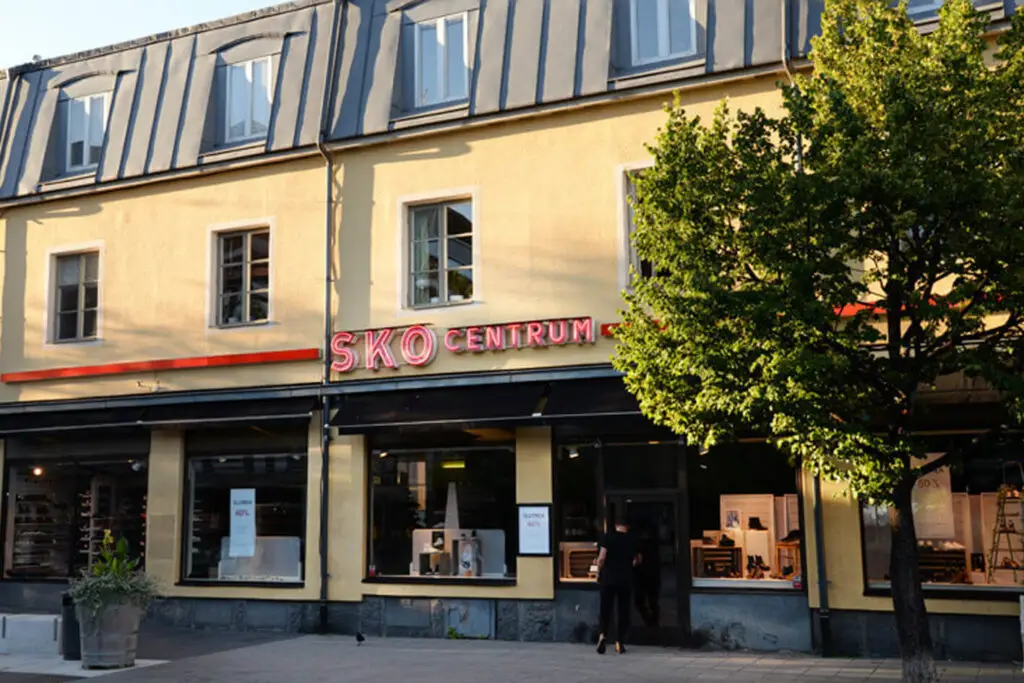 En gul byggnad med modern fasad, inrymmer "Sko Centrum" butik. Skyltfönstret har stora fönster som visar olika produkter. Ett träd på höger sida av stommen kastar en skugga på byggnaden. Några affischer och skyltar syns i fönstren.