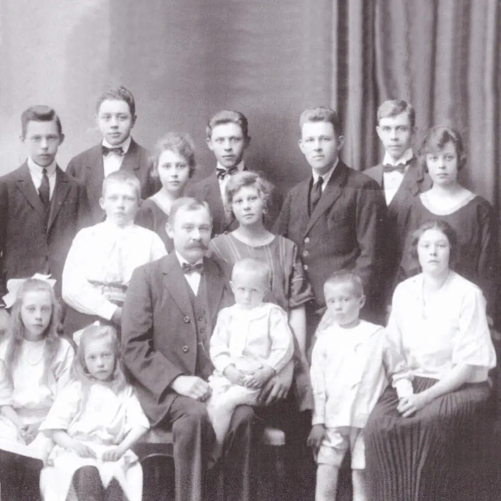 En vintage, svartvit fotografi med en stor familj. Bilden omfattar tre vuxna och elva barn i olika åldrar. Familjemedlemmarna är klädda i högtidskläder, de flesta står och några sitter framför. En gardin fungerar som bakgrund.